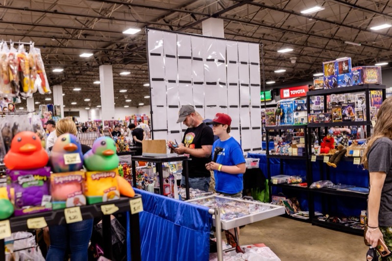 A busy aisle with a parent and child browsing shelves of toys under bright expo hall lights