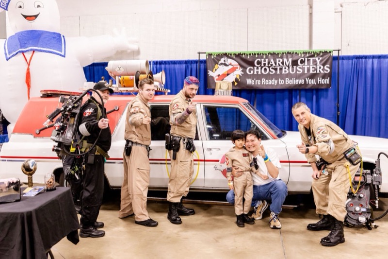 Charm City Ghostbusters posing with a young fan in a Ghostbusters jumpsuit beside the Ecto-1