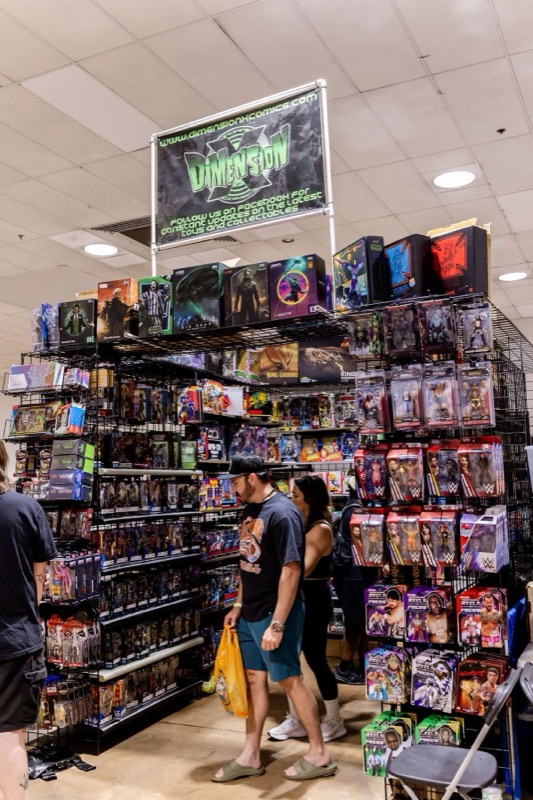 An attendee with a yellow shopping bag walking past a Dimension X booth packed with action figures