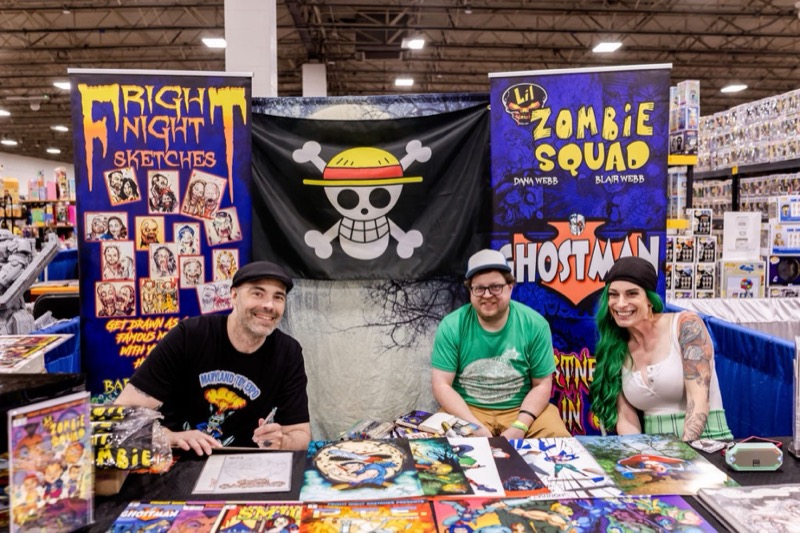 Three smiling independent artists at a booth of custom comics, prints, and horror art